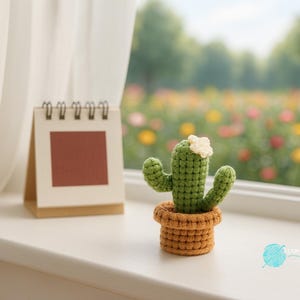 Puede incluir: Un cactus tejido a mano en una maceta marrón, con una pequeña flor blanca. Se encuentra en un alféizar blanco junto a un pequeño calendario. El fondo muestra una vista borrosa de un jardín de flores.