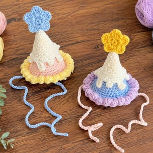 May include: Two crocheted party hats in pastel colors. Each hat features a cone shape with white frosting, a scalloped brim, and a star on top. One hat has a blue star and the other has a yellow star. Both have attached ties.