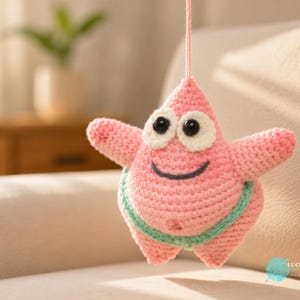 May include: A pink crochet starfish toy with a pointed top, two arms, and two legs. It features large black plastic eyes with white felt backing, a stitched dark blue smile, and a light green waistband. A pink yarn string is attached to the top.