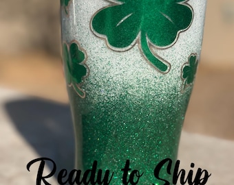 Puede incluir: Un vaso de glitter blanco y verde con un diseño de trébol de cuatro hojas. El vaso tiene un efecto degradado verde, que se desvanece del blanco al verde. "Ready to Ship" está escrito en texto negro en la parte inferior del vaso.