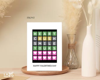 May include: A Valentine's Day greeting card with the words "HEART CRUSH SMILE YOURS" in a grid pattern. Below the grid are pink squares with white hearts. The card is on a wooden tray.