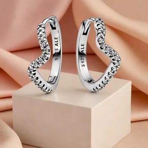 May include: A pair of silver heart-shaped hoop earrings. The earrings are encrusted with small, clear gemstones along the heart's curve. The inscription "S925 ALE" is visible on the inside of each earring. The earrings are displayed on a white cube.