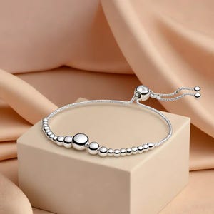 May include: A silver bracelet with a sliding clasp and a series of round beads. The bracelet features a central cluster of larger beads and smaller beads along the band. The background is a soft, neutral color.
