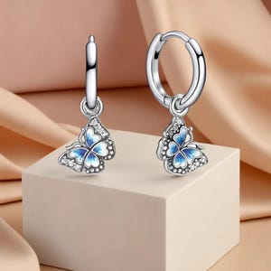 以下が含まれることがあります： 蝶のチャームが付いたシルバーのフープピアス。各チャームは、青と白の蝶のデザインとクリスタルのアクセントが特徴です。ピアスは、柔らかなピンク色の背景に対して、白い立方体に表示されています。