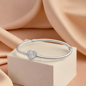 Könnte beinhalten: Ein silbernes Armband mit einem herzförmigen Anhänger, der mit kleinen, klaren Steinen besetzt ist. Das Armband wird auf einer weißen Oberfläche vor einem weichen, pfirsichfarbenen Hintergrund präsentiert. Das Armband hat ein strukturiertes, schlangenartiges Design.