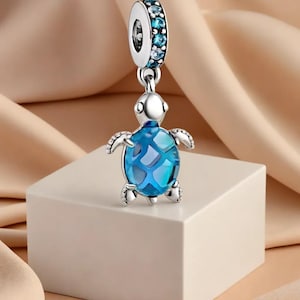Può includere: Un charm in argento con un design a forma di tartaruga blu. La tartaruga ha un corpo blu sfaccettato e piccole pinne dettagliate. Il charm è sospeso da un anello d'argento ornato di pietre blu. Il charm è esposto su una superficie bianca su uno sfondo color pesca.