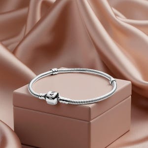 May include: A silver bracelet with a snake chain design and a square charm with the letters "PAN" on it. The bracelet is displayed on a small, square, light brown box against a backdrop of draped, peach-coloured fabric.