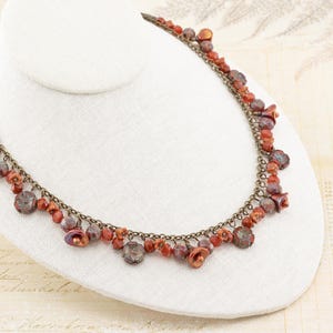 May include: A necklace with a bronze chain and various red and brown beads. The beads are in different shapes and sizes, including round, flower-shaped, and oblong. The necklace is displayed on a white bust.