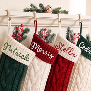 Könnte beinhalten: Vier Weihnachtsstrümpfe aus Zopfstrick, die an goldfarbenen Metallhaken hängen. Die Strümpfe haben cremefarbene Bündchen, die mit den Namen Patrcile, Morris, Stella und Cedric bestickt sind. Jeder Strumpf ist mit künstlichen Tannenzweigen und roten Beeren gefüllt.