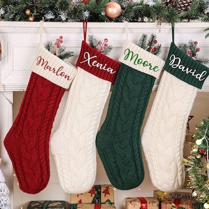 Könnte beinhalten: Vier Weihnachtssocken aus Zopfstrick hängen in einer Reihe. Die erste ist rot mit einer weißen Manschette, bestickt mit dem Namen Marlon. Die zweite ist weiß mit einer roten Manschette, bestickt mit Xenia. Die dritte ist grün mit einer weißen Manschette, bestickt mit Moore. Die vierte ist weiß mit einer grünen Manschette, bestickt mit David.