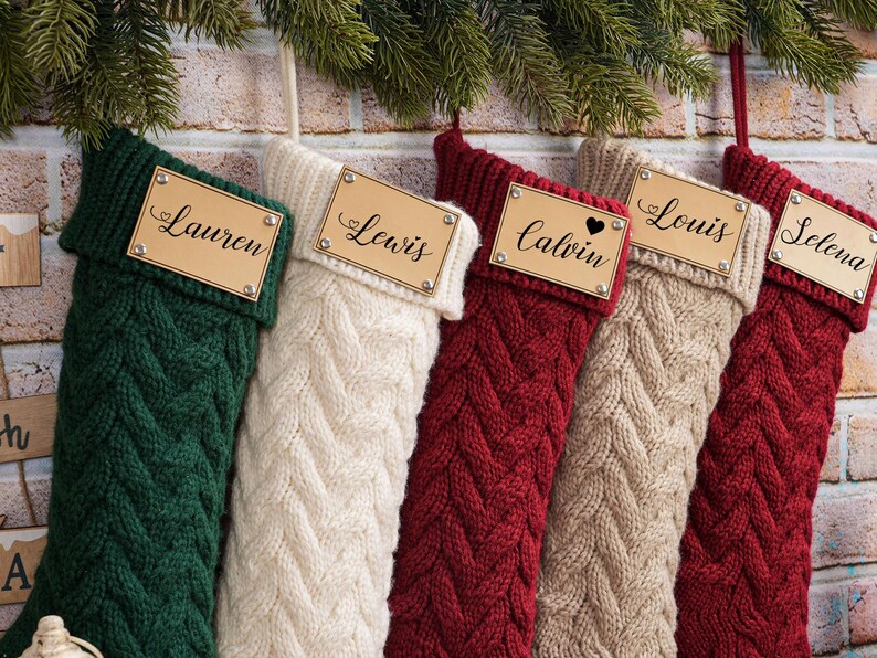 Puede incluir: Cinco calcetines navide&ntilde;os de punto trenzado en verde, crema, rojo, beige y burdeos, cada uno con una etiqueta de madera con un nombre. Nombres: Lauren, Lewis, Calvin, Louis y Selena. Decoraci&oacute;n festiva.