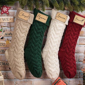 Puede incluir: Cuatro calcetines navide&ntilde;os de punto trenzado en beige, verde, blanco y rojo cuelgan de una pared de ladrillo. Cada calcet&iacute;n tiene una etiqueta de madera con un nombre: Chazel, Hyman, Lauren y Barrett. Decoraciones festivas, letrero de madera y casa de jengibre.