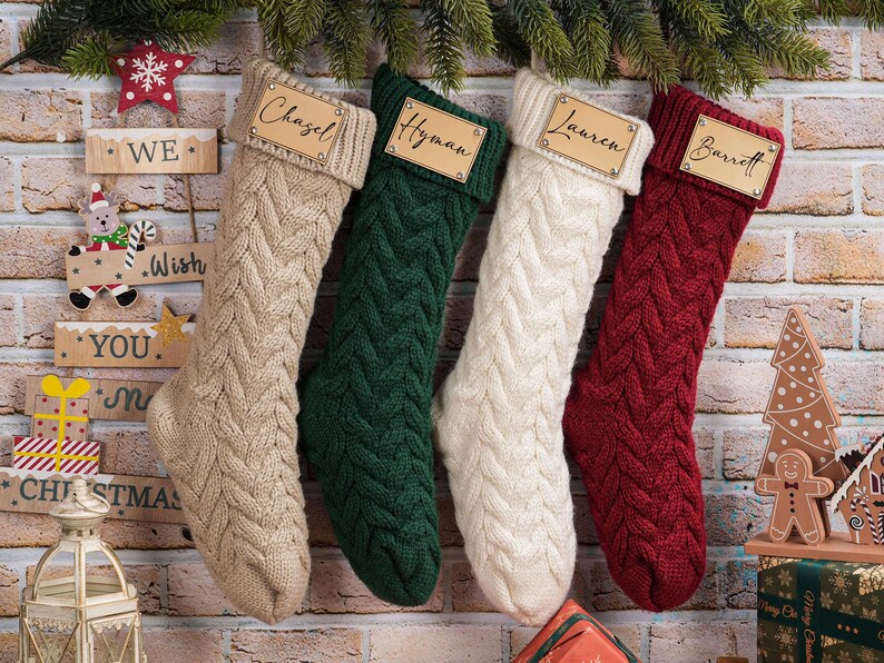 Puede incluir: Cuatro calcetines navide&ntilde;os de punto trenzado en beige, verde, blanco y rojo cuelgan de una pared de ladrillo. Cada calcet&iacute;n tiene una etiqueta de madera con un nombre: Chazel, Hyman, Lauren y Barrett. Decoraciones festivas, letrero de madera y casa de jengibre.