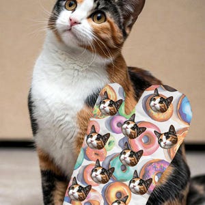 May include: A calico cat looks upwards, next to an oven mitt with a repeating pattern. The mitt design features cat faces inside donuts. The donuts are in various colours, including pink, blue, and yellow.