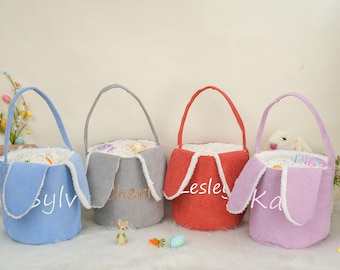May include: Four Easter baskets with bunny ears. The baskets are made of a soft, fuzzy material and are in the colors blue, gray, red, and purple. The baskets are decorated with the names "Sylv", "Cheri", "Lesles", and "Ka".