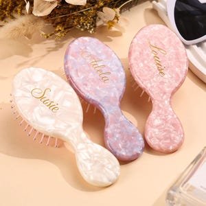 May include: Three oval hairbrushes with a marbled finish in shades of cream, lavender, and pink. Each brush features gold cursive text reading Susie, Adela, and Louise. The brushes have white bristles and are arranged side by side on a flat surface.