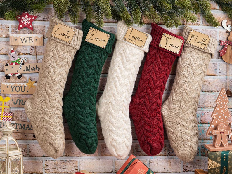 Puede incluir: Cinco calcetines navide&ntilde;os de punto trenzado en beige, verde, blanco, rojo y beige cuelgan de una pared de ladrillo. Cada calcet&iacute;n tiene una etiqueta de nombre de madera. Los nombres son Cynthia, Eunice, Lauren, Tyned y Martha. Ramas de pino verdes en la parte superior.