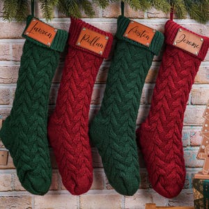 Puede incluir: Cuatro calcetines navide&ntilde;os de punto trenzado en verde y rojo cuelgan de una pared de ladrillo. Cada calcet&iacute;n tiene una etiqueta de cuero con un nombre en escritura. Decoraci&oacute;n festiva navide&ntilde;a.