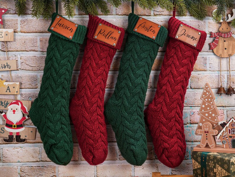 Puede incluir: Cuatro calcetines navide&ntilde;os de punto trenzado en verde y rojo cuelgan de una pared de ladrillo. Cada calcet&iacute;n tiene una etiqueta de cuero con un nombre en escritura. Decoraci&oacute;n festiva navide&ntilde;a.