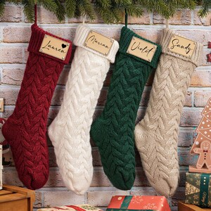 Puede incluir: Cuatro calcetines navide&ntilde;os de punto trenzado en rojo, blanco, verde y beige cuelgan de una pared de ladrillo. Cada calcet&iacute;n tiene una etiqueta de nombre de madera. Elementos decorativos navide&ntilde;os: un reno de madera, una casa de jengibre y regalos envueltos.