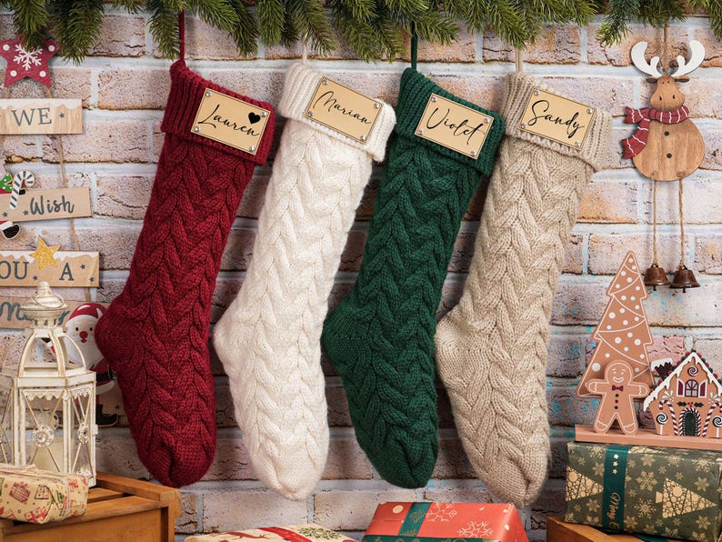 Puede incluir: Cuatro calcetines navide&ntilde;os de punto trenzado en rojo, blanco, verde y beige cuelgan de una pared de ladrillo. Cada calcet&iacute;n tiene una etiqueta de nombre de madera. Elementos decorativos navide&ntilde;os: un reno de madera, una casa de jengibre y regalos envueltos.
