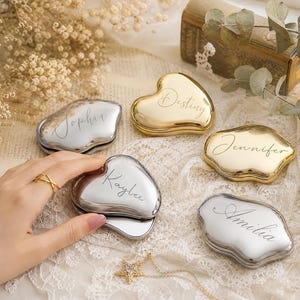 Peut inclure: Une collection de miroirs de poche en forme de cœur avec des finitions métalliques dorées et argentées. Chaque miroir présente un prénom personnalisé gravé en écriture cursive, incluant Sophia, Destiny, Jennifer, Kaylee et Amilia.