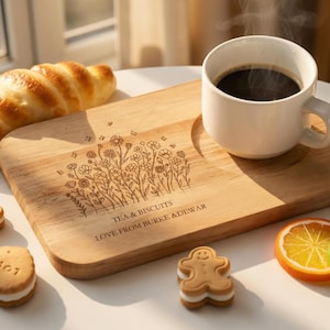 Op de afbeelding: Een houten serveerplank met een bloemenontwerp en de tekst "TEA & BISCUITS LOVE FROM BURKE & DEWAR" bevat een witte koffiekop, een croissant en koekjes. Gesneden citrusvruchten en madeliefjes zijn ook aanwezig.