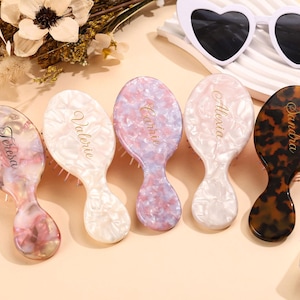 May include: Five oval hairbrushes with marbled patterns in pink, white, lavender, and tortoiseshell. Each brush features a personalized name in gold script: Teresa, Valerie, Carrie, Alexia, and Sandra. The brushes have white bristles.
