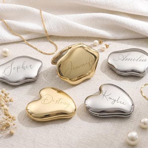 Peut inclure: Cinq miroirs de poche en métal poli aux tons or et argent, chacun présentant une forme organique irrégulière et un nom gravé personnalisé dont Sophia, Jennifer, Amilia, Destiny et Kaylee, disposés avec des chaînes dorées et des perles blanches.