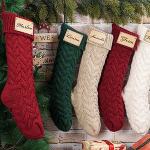 Puede incluir: Cinco calcetines navide&ntilde;os de punto trenzado en rojo, verde, crema y blanco cuelgan de una pared de ladrillo. Cada calcet&iacute;n tiene una etiqueta de nombre de madera. Los nombres en las etiquetas incluyen Marlin, Lauren, Amanda, Gloria y Service. Decoraci&oacute;n festiva navide&ntilde;a.