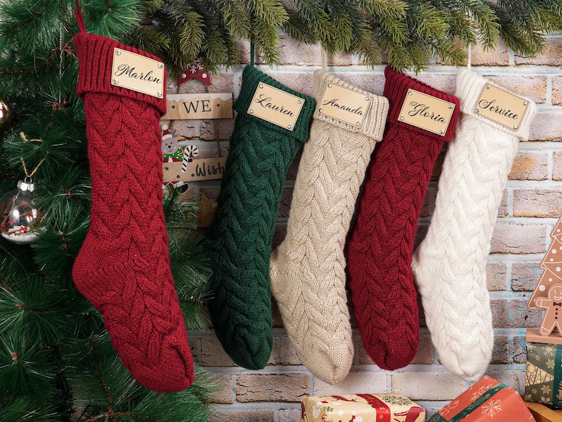Puede incluir: Cinco calcetines navide&ntilde;os de punto trenzado en rojo, verde, crema y blanco cuelgan de una pared de ladrillo. Cada calcet&iacute;n tiene una etiqueta de nombre de madera. Los nombres en las etiquetas incluyen Marlin, Lauren, Amanda, Gloria y Service. Decoraci&oacute;n festiva navide&ntilde;a.