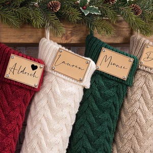 May include: Four cable-knit Christmas stockings in red, cream, dark green, and beige. Each stocking features a rectangular leather-like name tag attached with four metal rivets. The tags are engraved with the names Aldrich, Lauren, Minnie, and Derrick.