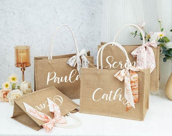 May include: Four personalized burlap tote bags with white lettering and colorful fabric bows. The bags are labeled "Paula", "Cath", "Scri", and "Sur".