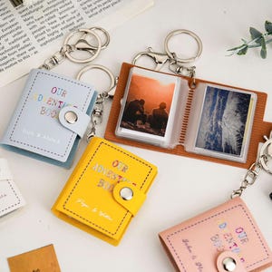 May include: A collection of small photo album keyrings in shades of blue, yellow, and pink. Each keyring has a miniature book design with the words "Our Adventure Book" and a snap closure. Some albums contain photos.