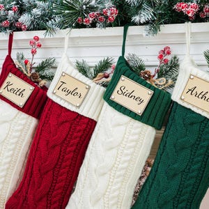 May include: Four cable-knit Christmas stockings in red, green, and white hang from a mantel. Each stocking has a wooden name tag that reads "Keith", "Taylor", "Sidney", and "Arthur". The mantel is decorated with greenery and red berries.
