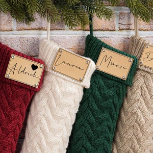 Könnte beinhalten: Vier Zopfstrick-Weihnachtsstrümpfe in Rot, Weiß, Grün und Beige hängen vor einer Backsteinmauer. Jeder Strumpf hat ein Namensschild aus Holz mit einem in Schreibschrift geschriebenen Namen. Der rote Strumpf hat ein Herzsymbol.