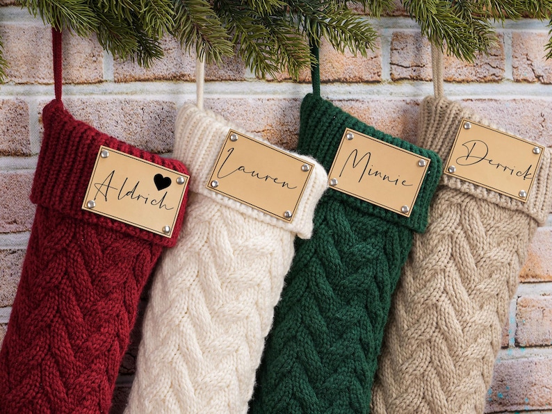 Puede incluir: Cuatro calcetines navide&ntilde;os de punto trenzado en rojo, blanco, verde y beige cuelgan contra una pared de ladrillos. Cada calcet&iacute;n tiene una etiqueta de nombre de madera con un nombre escrito en cursiva. El calcet&iacute;n rojo tiene un s&iacute;mbolo de coraz&oacute;n.