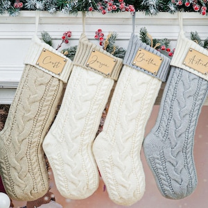 May include: Four cable-knit Christmas stockings in beige, white, and gray hang from a mantel. Each stocking has a wooden name tag: Croft, Joyce, Bertha, and Astrid. A wooden reindeer ornament and greenery decorate the mantel.