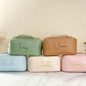 Puede incluir: Seis neceseres personalizados en colores pastel: verde, beige, azul claro, crema y rosa. Cada neceser tiene un asa y un nombre en letra cursiva. Los neceseres están colocados sobre una superficie de mármol.