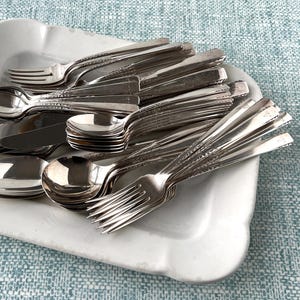 May include: A collection of vintage silverware, including forks, spoons, and knives, arranged on a white, scalloped-edge serving dish. The silverware has a shiny, reflective surface, and the dish sits on a light blue patterned surface.