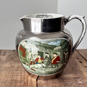 Peut inclure: Pichet argenté avec une scène historique peinte à la main. L'œuvre représente des personnages en costume d'époque, sur fond de bâtiments et d'un chien. Le bord du pichet porte l'inscription "Death of..."