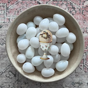 May include: A beige bowl filled with white, egg-shaped stones. A decorative egg, featuring a chick hatching, sits in a floral egg cup. The egg cup has a gold base and rim. The text on the egg reads "All Easter Greetings".