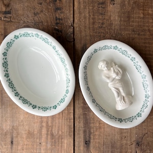 May include: Two oval, white ceramic dishes with a green floral border. One dish is empty, the other contains a small white figurine of a woman. The dishes are on a rustic, wooden surface.