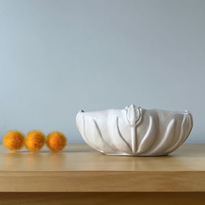 May include: A cream-colored, decorative ceramic bowl with a floral design. Three small, orange, fuzzy balls are arranged to the left of the bowl on a wooden surface. The background is a light blue wall.