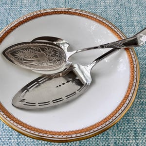 May include: Two silver serving utensils rest on a white plate with an orange and gold rim. One utensil has a decorative, oval-shaped head with an etched design. The other has a slotted, rectangular head. The plate is on a blue patterned surface.