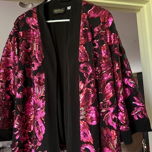 May include: A black open-front jacket with vibrant pink sequin floral patterns. The jacket has a black collar and trim, and is hanging on a white hanger. The sequins create a shimmering effect, adding a touch of glamour to the garment.