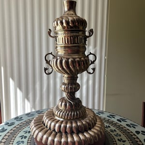 Puede incluir: Un objeto decorativo de latón alto y ornamentado con una base escalonada y un cuerpo bulboso acanalado. Cuenta con asas decorativas y un remate puntiagudo. El objeto se exhibe sobre una mesa redonda con un patrón de mosaico.