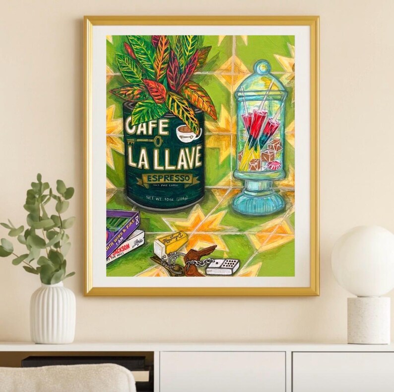 May include: A framed art print featuring a green Cafe La Llave espresso can with a plant, a glass apothecary jar filled with red and yellow popsicles, stacked books, a small yellow box, a brown key, and a white domino on a yellow star tile floor.