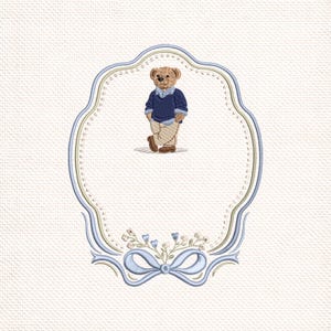 May include: Embroidered design of a teddy bear in a blue sweater and beige trousers, within a decorative oval frame. The frame is outlined in blue and gold, with a bow and floral details at the base. The background has a textured, off-white appearance.