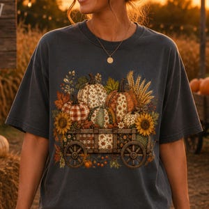 Puede incluir: Una camiseta gris carbón de manga corta con un estampado gráfico de un carro de madera lleno de calabazas con varios patrones, girasoles y tallos de trigo. El diseño incluye hojas de otoño y follaje centrado en el pecho de la prenda.